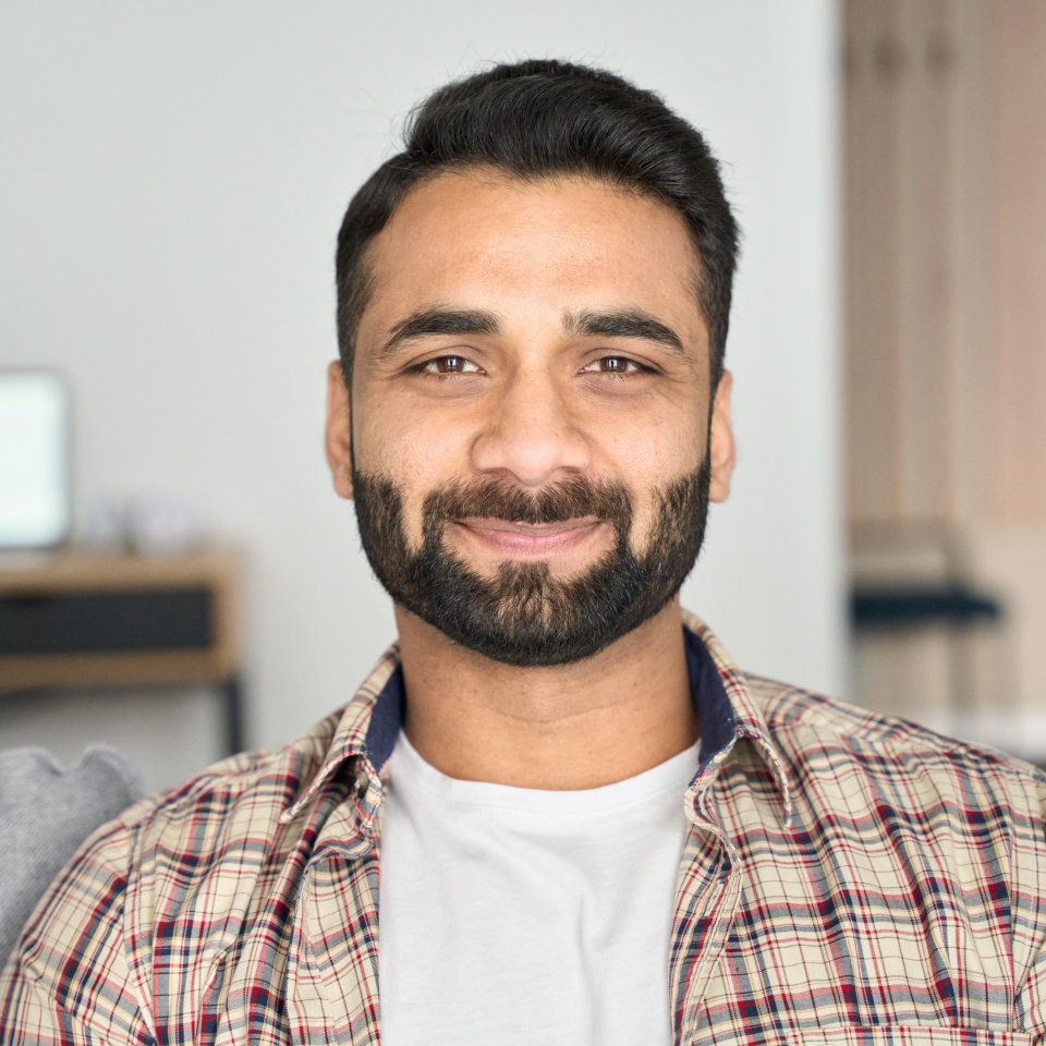 Headshot portrait of attractive confident indian Hispanic man looking at camera sitting on couch at modern living room. Latin businessman posing in casual stylish look at home office.