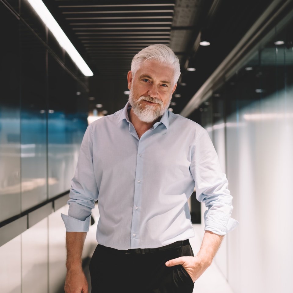 Handsome elderly man in elegant shirt looking at camera and keeping hand in pocket while standing near glass wall of modern corridor