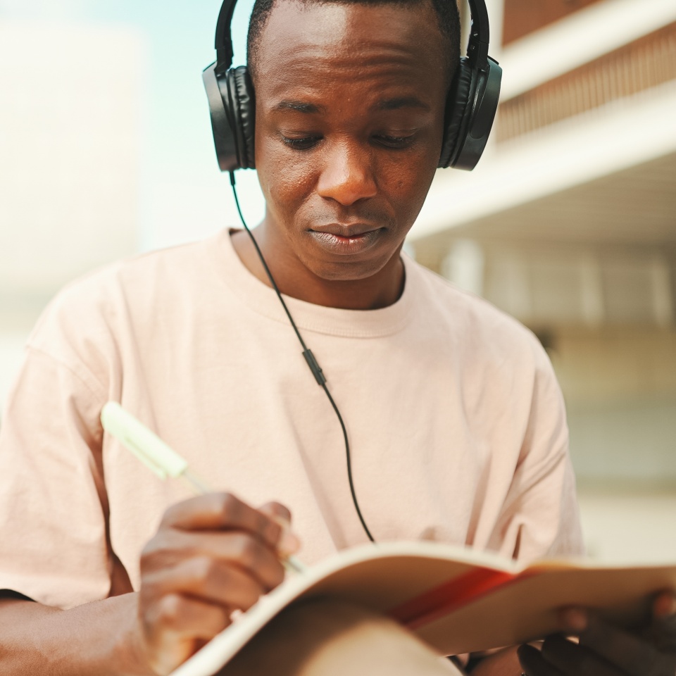 Young african student sitting outside of university and taking notes while study online with headphones