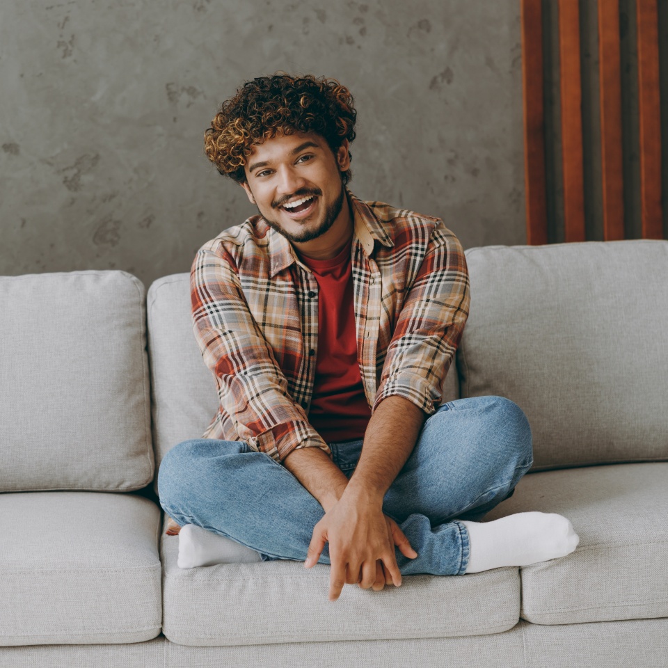 Full body smiling happy fun young Indian man wears casual clothes looking camera sits on grey sofa couch stay at home hotel flat rest relax spend free spare time in living room indoor. Lounge concept