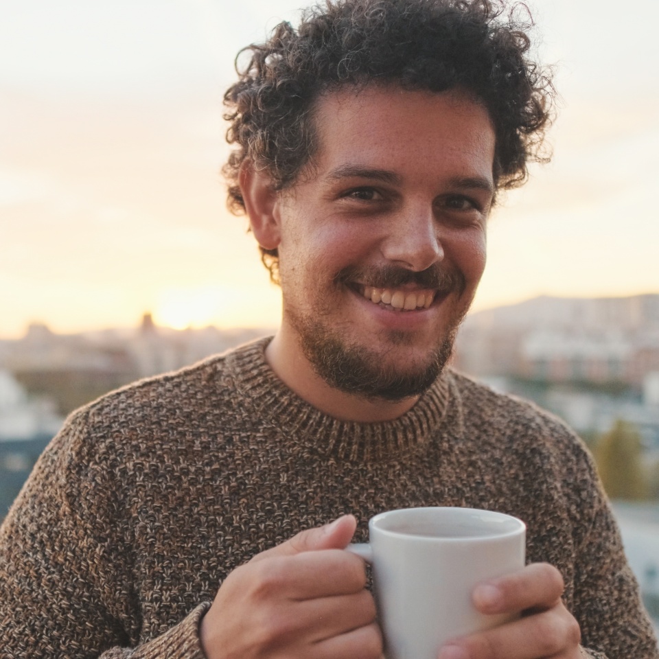 Peaceful guy with a cup of coffee stands on the balcony looks at camera and smiling