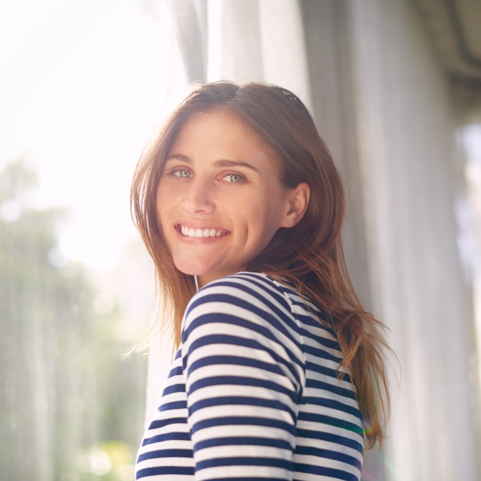 Woman, smile and portrait in house for relax and calm for holiday and cheerful on blurred background. Young person and happy with window for view of garden or backyard with sunlight in lounge