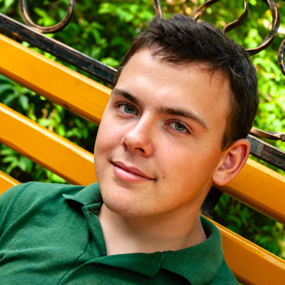 Young caucasian male relaxing on bright yellow bench in a lush green park setting, wearing a green shirt and smiling with contentment.