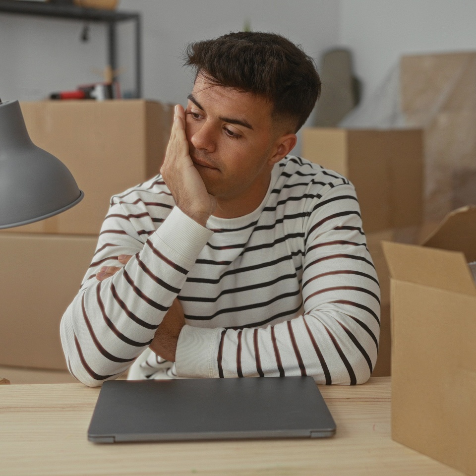 Young man frustrated in cluttered apartment with moving boxes and laptop, sitting at table under lamp with hand on forehead in modern indoor setting