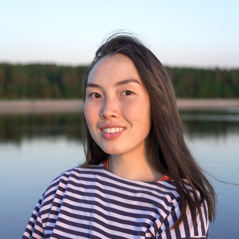 A woman in a striped shirt smiles while standing near a calm lake at sunset, with forest reflecting in the water.