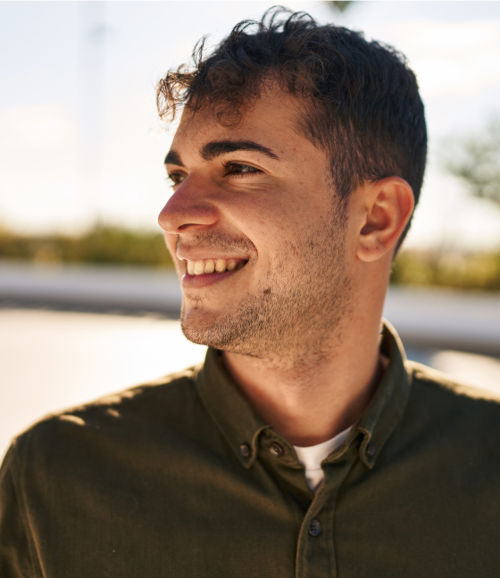 A young man with short curly hair and stubble, wearing a button-up shirt, smiles while looking to the side outdoors in natural light.