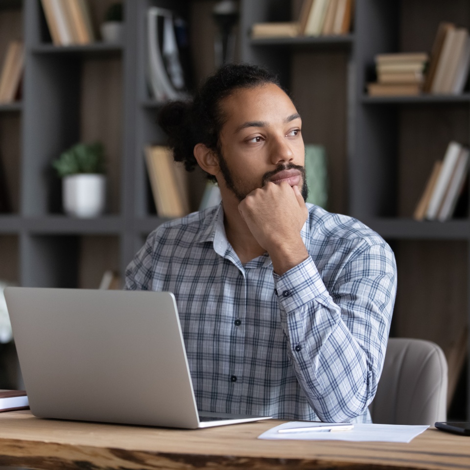 Serious thoughtful young African entrepreneur working at laptop at home workplace table, thinking over business issues solving, online startup project, making decision, looking away