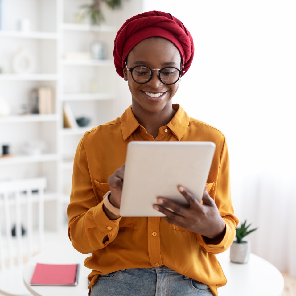Pretty stylish african american woman in casual outfit standing by worktable, using digital tablet and smiling, posing alone at cozy office, copy space. Gadgets and business concept