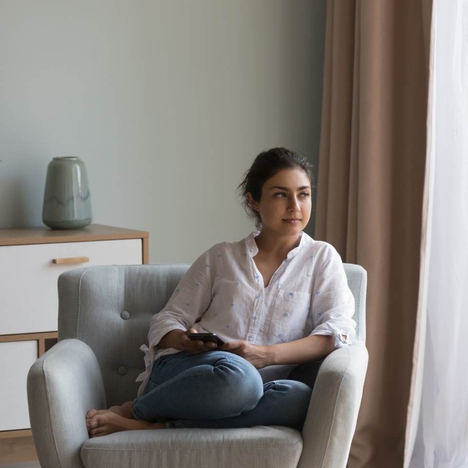 Attractive pensive Indian woman resting on cozy armchair staring into distance spend weekend leisure alone at modern home. Thoughtful female thinking seated on cozy chair looking peaceful and serious