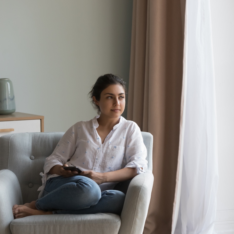 Attractive pensive Indian woman resting on cozy armchair staring into distance spend weekend leisure alone at modern home. Thoughtful female thinking seated on cozy chair looking peaceful and serious