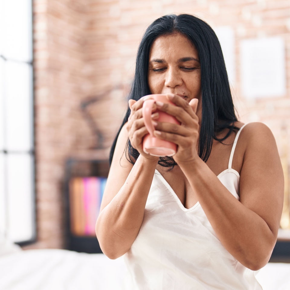 Middle age hispanic woman drinking cup of coffee sitting on bed at bedroom