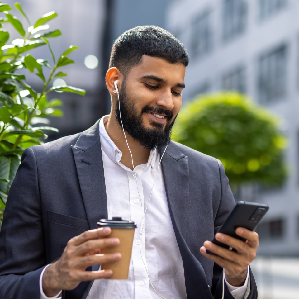 A cheerful young businessman wearing a suit is enjoying music with headphones and holding a coffee cup while using a smartphone ,outside an office building surrounded by greenery.