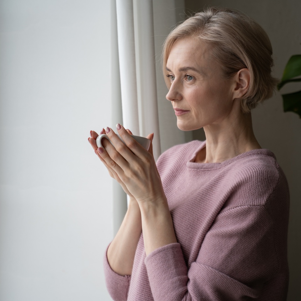 A woman stands by the window, holding a warm cup of tea in her hands. She gazes outside, lost in thought, with a look of tranquility on her face. The soft natural light highlights the peaceful mood of