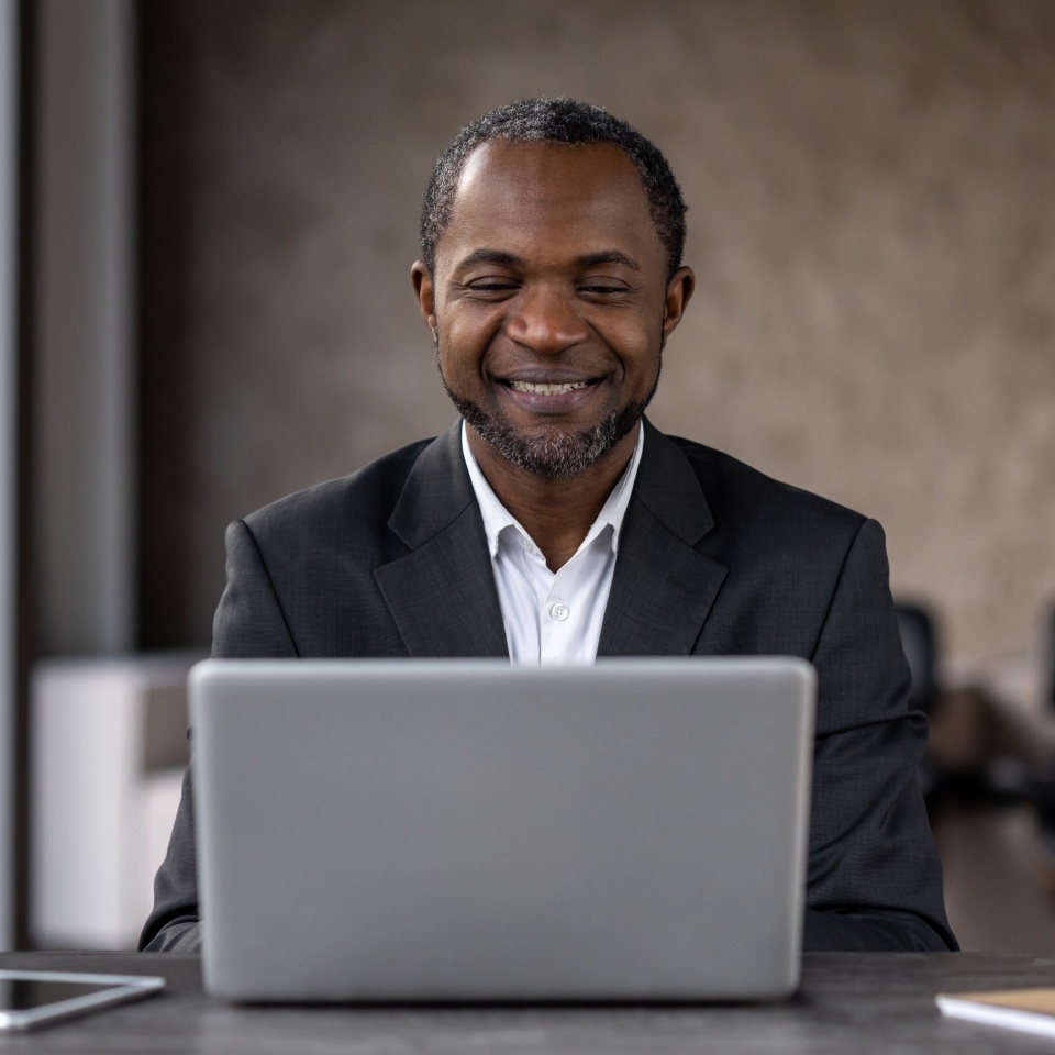 A professional African American man smiles while working on his laptop in an office setting. This image captures a moment of career satisfaction and confidence.