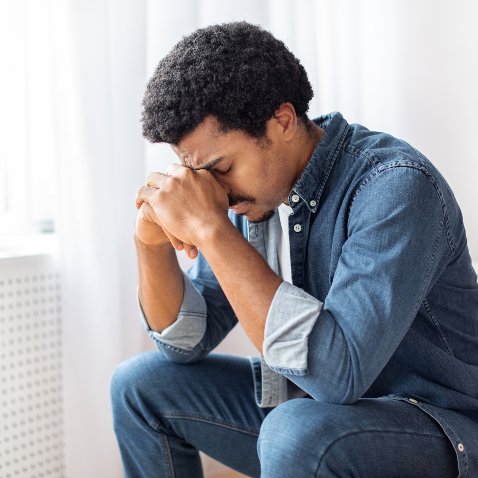 Black man is seated on a couch, with one hand pressed against his face in a contemplative gesture. He appears deep in thought, possibly experiencing emotions like stress, sadness, or exhaustion.