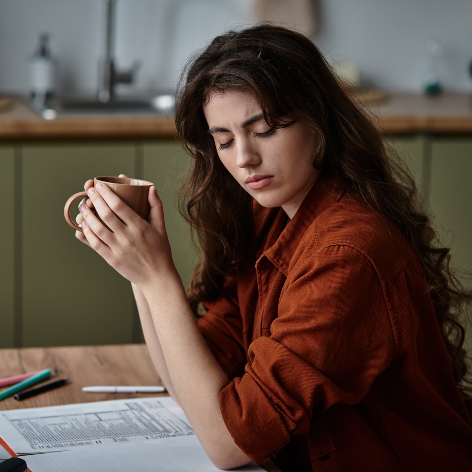 A woman sits at a table, holding a cup while contemplating her thoughts in a cozy kitchen.
