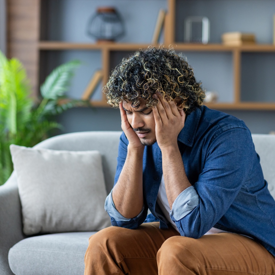 Stressed man with curly hair sitting on a couch, experiencing a bad headache in a modern interior. He is overwhelmed with negative emotions.