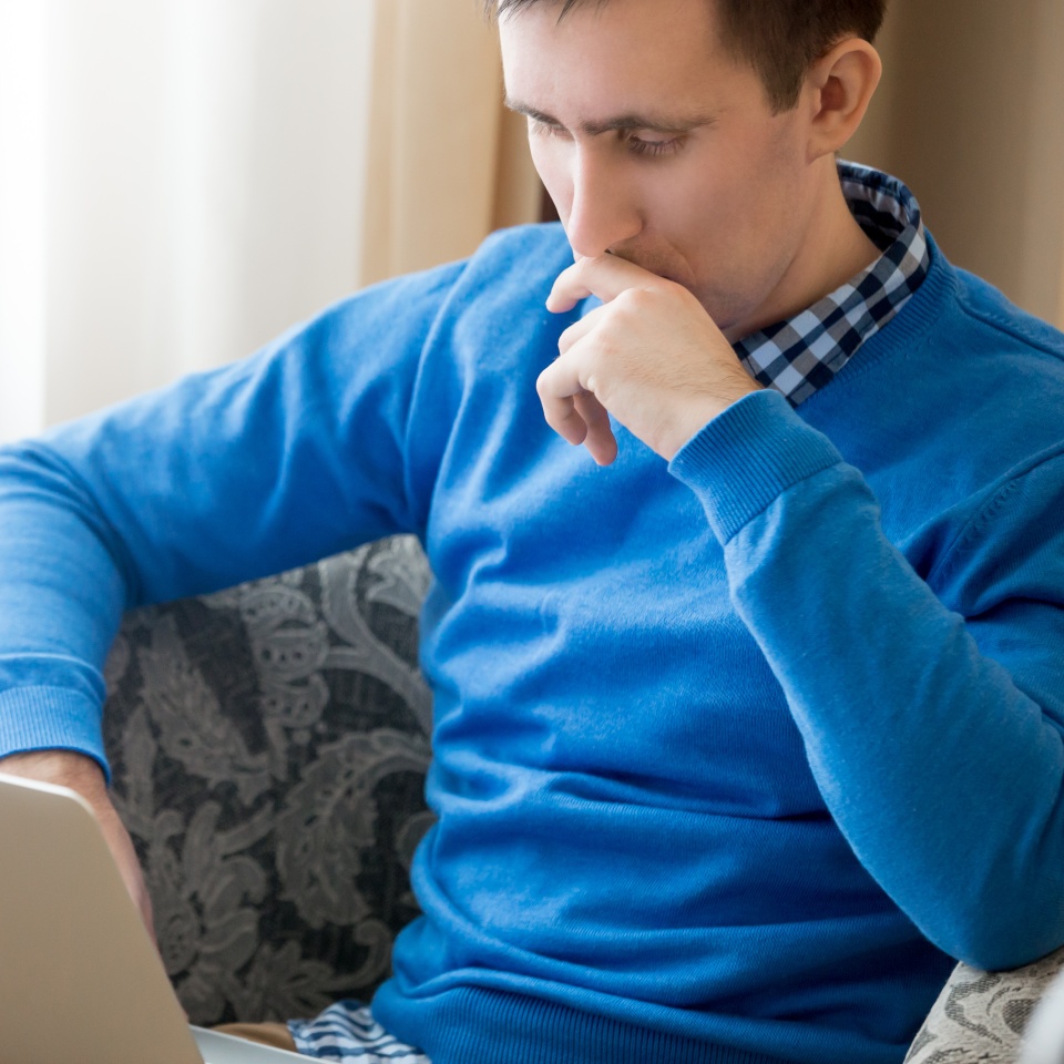Portrait of pensive casual attractive young man sitting in armchair indoors, concentrated on work, male model using laptop, looking at the screen with serious thoughtful expression