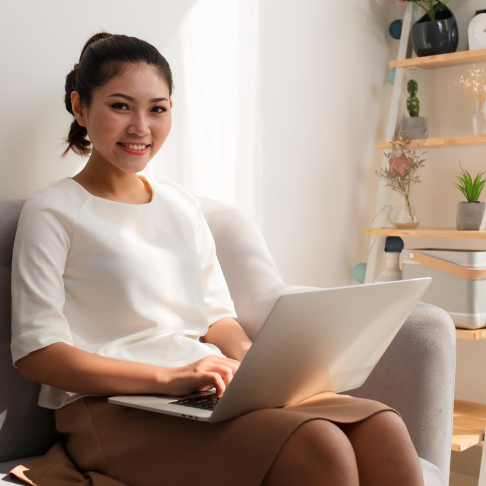 Woman sitting on a sofa using a laptop, smiling at the camera. Sunlight enters from the left, and a shelf with plants and decor is visible in the background.