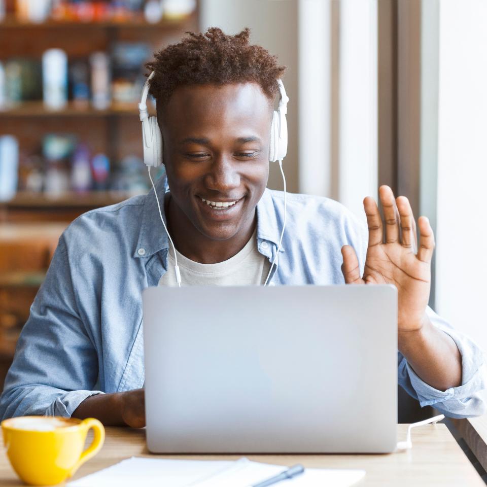 A person wearing headphones sits at a desk, smiling and waving at a laptop screen, with a yellow mug and notebook nearby.