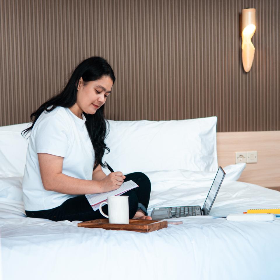 A woman sits on a bed, writing in a notebook with a pen. A laptop, mug, and books are on the bed beside her.