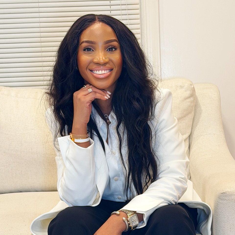 A woman with long wavy hair, wearing a white blazer and black pants, sits on a light-colored sofa and smiles at the camera.